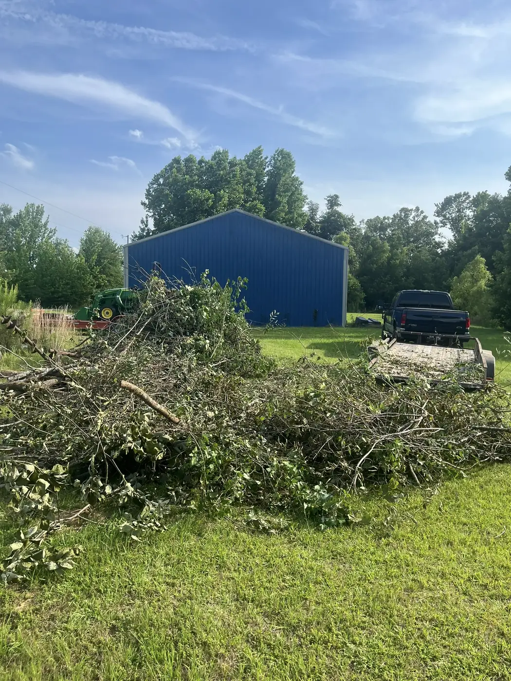 Truck bed and trailer piled with brush and tree branches in a rural yard near a blue metal barn